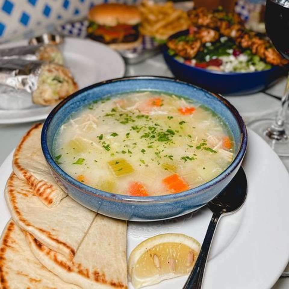 A bowl of chicken and vegetable soup with pita bread and a lemon wedge, alongside other dishes.