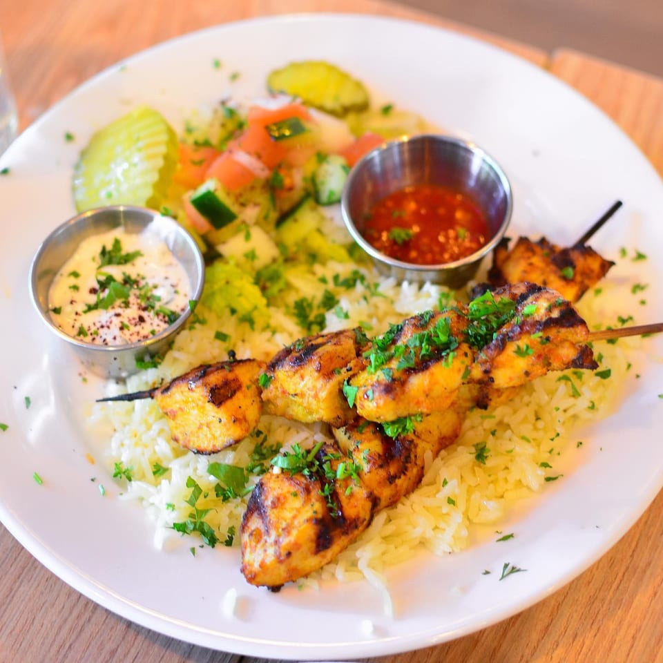 Chicken kebab skewers with rice, salad, tzatziki, and chili sauce on a white plate.