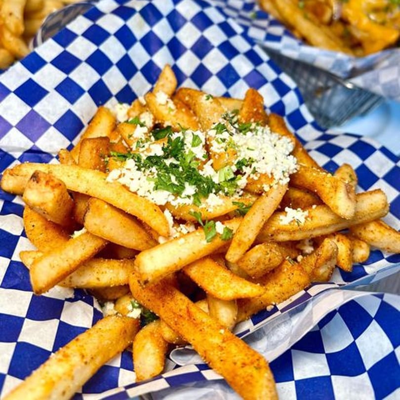 Seasoned fries topped with crumbled feta cheese and fresh parsley in a blue and white checkered basket.